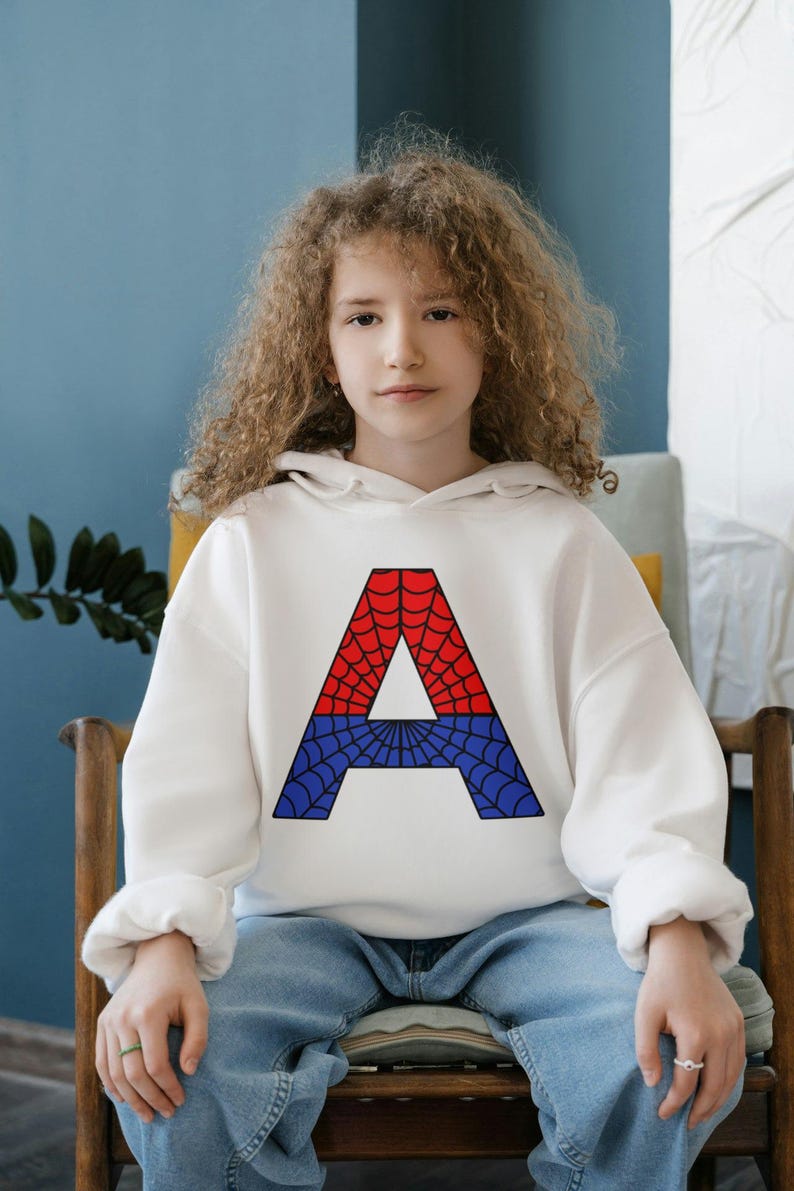May include: A white hoodie with a red and blue spiderweb letter A on the front. The hoodie is worn by a young person with curly hair sitting on a wooden chair.