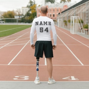 May include: A person wearing a white long-sleeved shirt with the text "NAME 54" on the back. They are wearing black shorts and a prosthetic leg. The person is standing on a red running track.