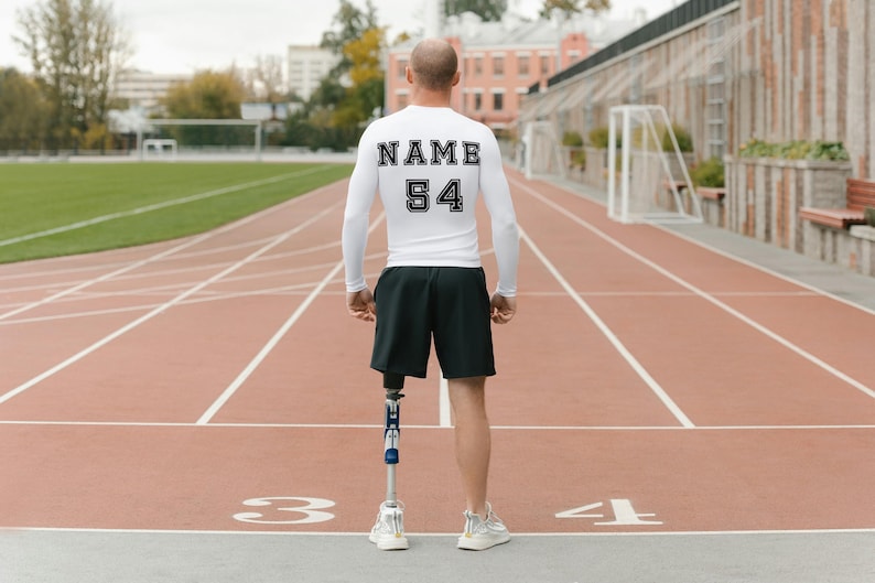 May include: A person wearing a white long-sleeved shirt with the text "NAME 54" on the back. They are wearing black shorts and a prosthetic leg. The person is standing on a red running track.