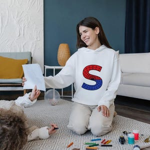 May include: A woman wearing a white hoodie with a red and blue letter "S" on the front is kneeling on a carpet. She is looking at a child who is lying on the carpet and drawing with a crayon. There are crayons and paint bottles on the carpet.