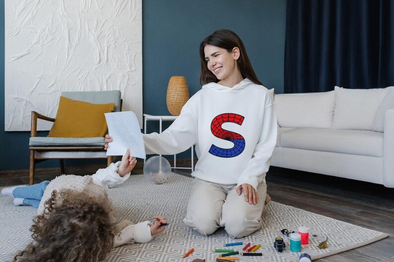 May include: A woman wearing a white hoodie with a red and blue letter "S" on the front is kneeling on a carpet. She is looking at a child who is lying on the carpet and drawing with a crayon. There are crayons and paint bottles on the carpet.