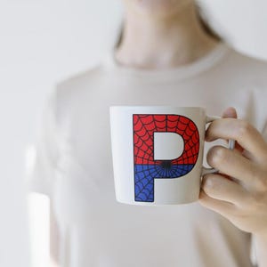 May include: A white ceramic mug with a red and blue spiderweb design featuring the letter "P" in the center.
