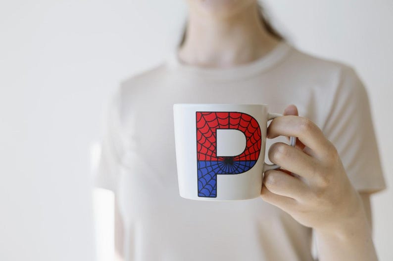 May include: A white ceramic mug with a red and blue spiderweb design featuring the letter "P" in the center.