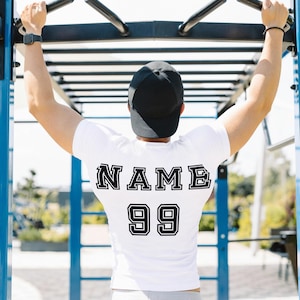 May include: A white t-shirt with black text on the back that says "NAME 99". The person is wearing a black baseball cap and is doing a pull-up on a blue and black exercise bar.