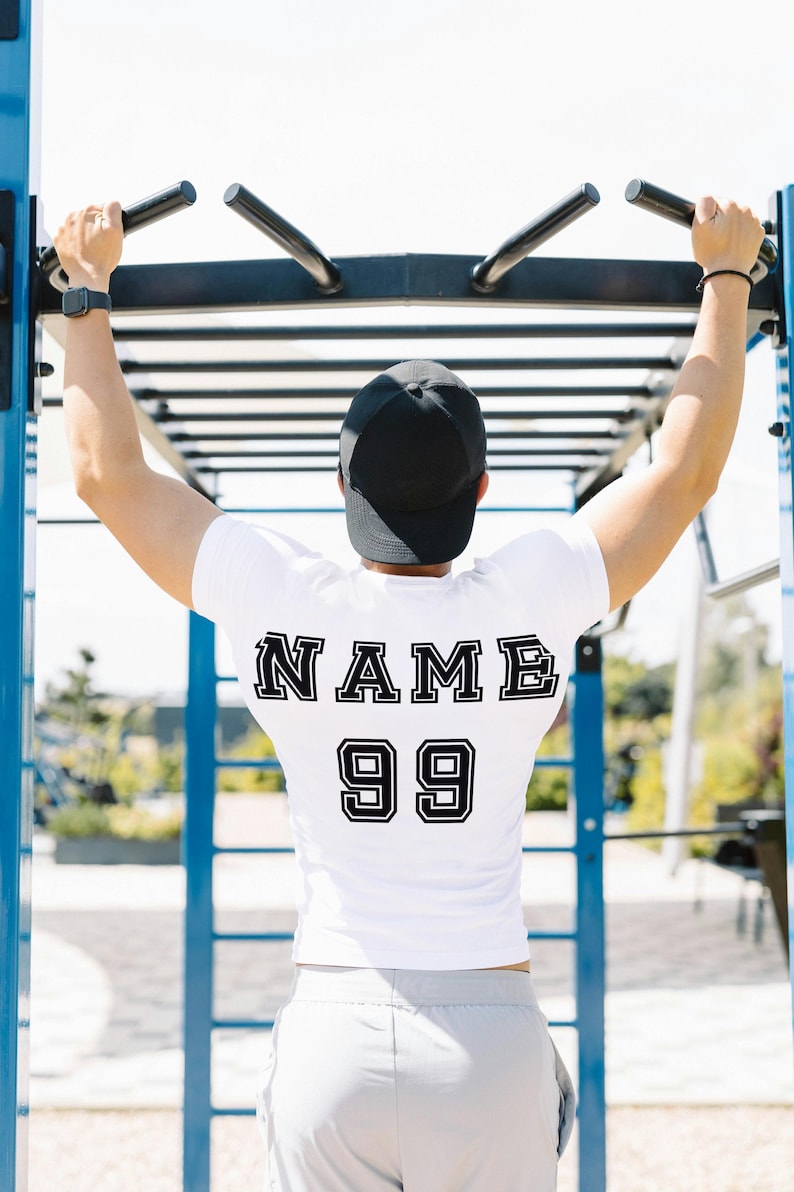 May include: A white t-shirt with black text on the back that says "NAME 99". The person is wearing a black baseball cap and is doing a pull-up on a blue and black exercise bar.