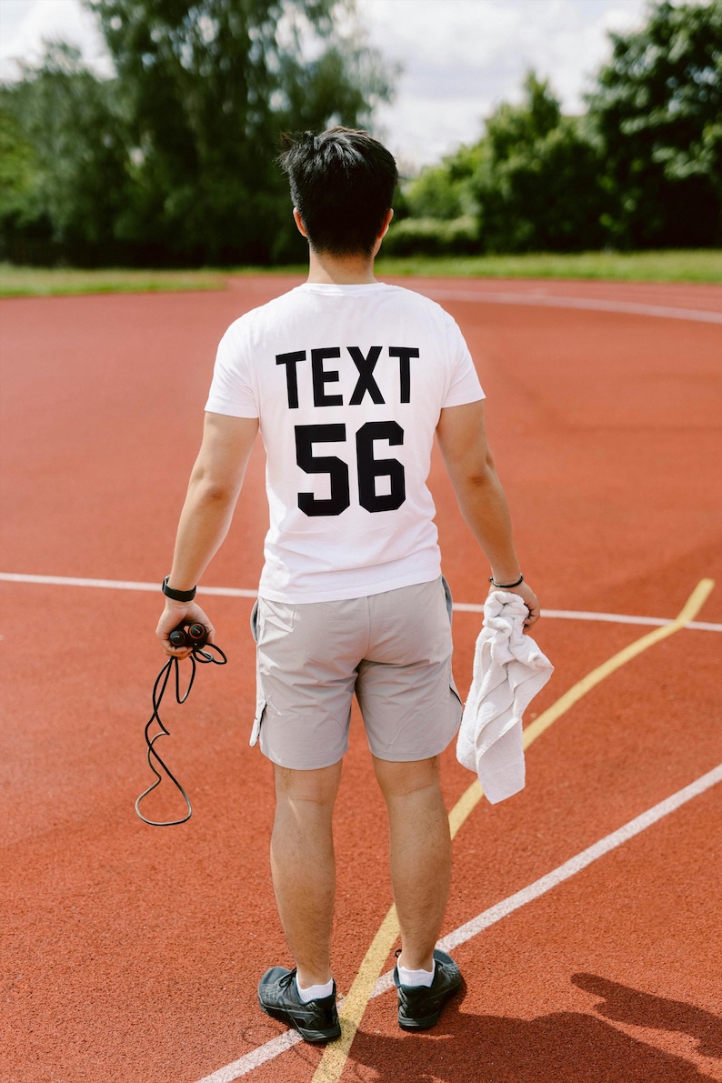 May include: A person wearing a white t-shirt with the text "TEXT 56" printed on the back. They are standing on a red track and holding a jump rope.
