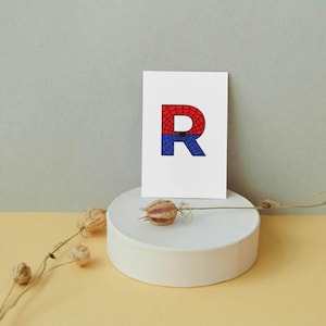 May include: A white card with a red and blue letter R on it. The letter R is filled with a pattern that resembles a spider web.