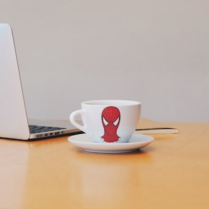 May include: A white coffee cup with a red and black Spider-Man design sits on a saucer on a wooden table. A laptop computer is in the background.
