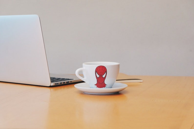 May include: A white coffee cup with a red and black Spider-Man design sits on a saucer on a wooden table. A laptop computer is in the background.