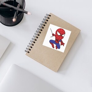 May include: A brown notebook with a white sticker featuring a cartoon illustration of Spiderman in a red and blue suit, swinging from a web.