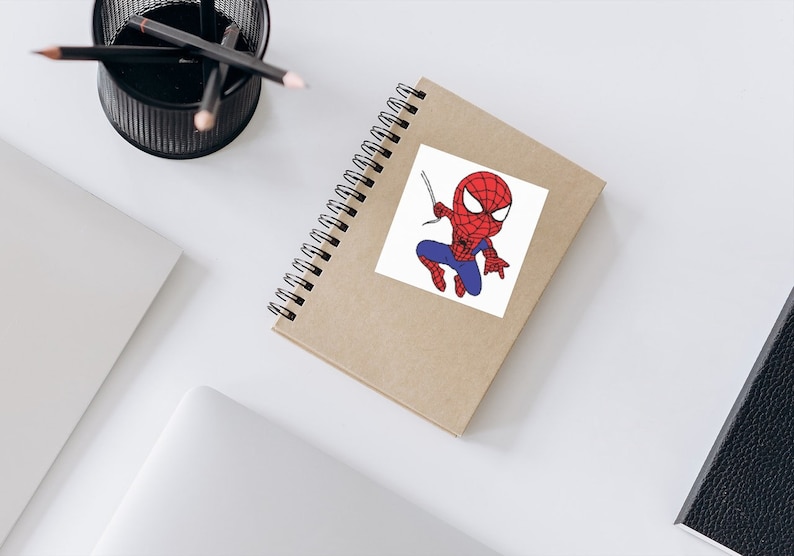 May include: A brown notebook with a white sticker featuring a cartoon illustration of Spiderman in a red and blue suit, swinging from a web.