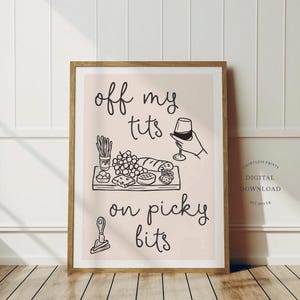 Könnte beinhalten: Ein gerahmter Kunstdruck mit dem Text "off my tits on picky bits" in handschriftlichem Stil. Das Bild zeigt eine Charcuterie-Platte mit Trauben, Käse und Brot sowie eine Hand, die ein Weinglas hält. Der Druck ist in einem Holzrahmen.