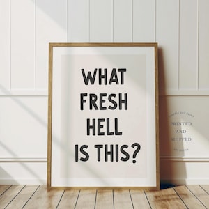 Op de afbeelding: Een ingelijst print met de tekst "What Fresh Hell Is This?" in zwarte letters op een witte achtergrond.