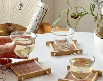 May include: Clear glass teacups with a black tree design, filled with tea, sit on bamboo coasters. A hand holds a teacup. A fan and scroll are in the background, suggesting a tea ceremony.