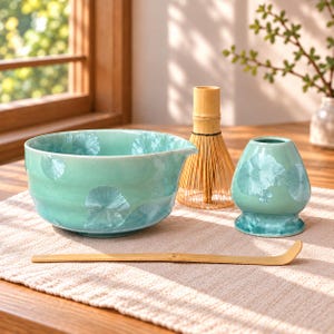May include: A light turquoise ceramic matcha bowl with a crystalline glaze pattern, a bamboo whisk, a bamboo tea scoop, and a matching turquoise ceramic whisk holder. The bowl features a small pouring spout on one side and a smooth, rounded shape.