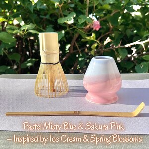 May include: A bamboo matcha whisk, a ceramic whisk holder with a white top and pink base, and a long bamboo scoop. The text reads: Pastel Misty Blue & Sakura Pink &ndash; Inspired by Ice Cream & Spring Blossoms. The items are arranged on a light surface.