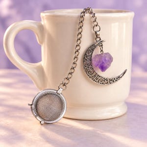 May include: A silver-toned metal tea infuser featuring a round mesh basket attached to a chain. The chain connects to a decorative crescent moon charm with intricate filigree cutouts and a small, rough-cut purple amethyst crystal dangling from it.