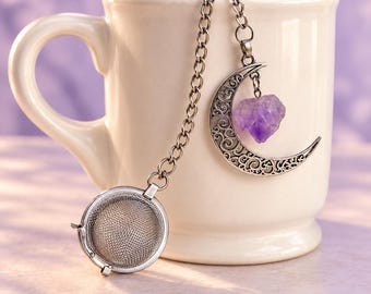 May include: A silver-toned metal tea infuser featuring a round mesh basket attached to a chain. The chain connects to a decorative crescent moon charm with intricate filigree cutouts and a small, rough-cut purple amethyst crystal dangling from it.