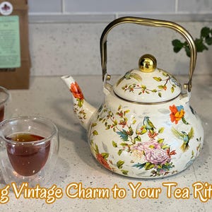 Pu&ograve; includere: Una teiera in smalto bianco con manico e pomello dorati, ornata da motivi floreali e di uccelli. Due tazze di t&egrave; in vetro trasparente sono davanti alla teiera. Il testo "Bring Vintage Charm to Your Tea Rituals" &egrave; in basso.