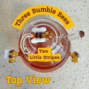 May include: A glass honey pot with three bumble bee decorations. The honey pot has a glass straw and the text "Three Bumble Bees", "Two Little Stripes", and "Top View". The honey pot is filled with honey.