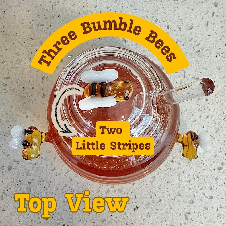 May include: A glass honey pot with three bumble bee decorations. The honey pot has a glass straw and the text "Three Bumble Bees", "Two Little Stripes", and "Top View". The honey pot is filled with honey.