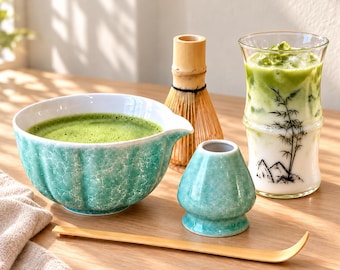 May include: A matcha tea set including a turquoise crackle-glaze bowl filled with green tea, a bamboo whisk, a matching turquoise whisk holder, a bamboo scoop, and a glass cup with a bamboo and mountain print containing an iced matcha latte.