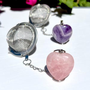 Könnte beinhalten: Zwei silberne Tee-Infuser mit herzförmigen Amethyst- und Rosenquarzsteinen, die an Ketten befestigt sind. Die Infuser sind zum Aufgießen von losen Blatttee konzipiert.