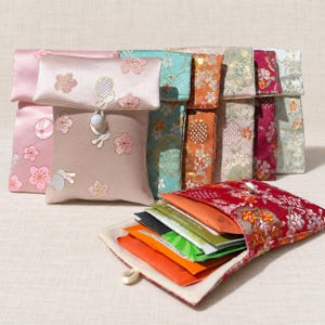 May include: A collection of rectangular fabric pouches featuring embroidered floral patterns in various colors including pink, teal, orange, and red. Each pouch has a flap closure secured with a white bead button. One red pouch is open, revealing several colorful tea bags inside.