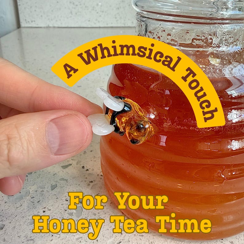 May include: A glass honey jar with a decorative bee accent. The bee has white wings and a black and amber body. The text "A Whimsical Touch" and "For Your Honey Tea Time" are displayed in yellow.