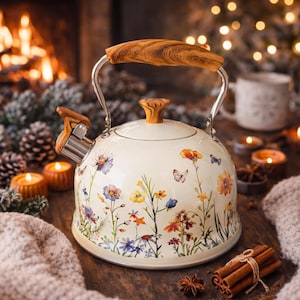 Könnte beinhalten: Cremefarbene Emaille-Teekanne mit floralem Design aus Wildblumen und Schmetterlingen. Sie hat einen Holzgriff, einen Ausgusshebel und einen Knauf. Die Kanne steht auf einer Holzoberfläche. Das Design umfasst eine Vielzahl bunter Blumen.