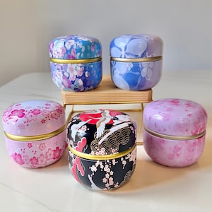 Puede incluir: Cinco latas decorativas de metal con tapas. Las latas presentan diseños florales e inspirados en Japón en tonos de azul, rosa y negro. Cada lata tiene una banda dorada en el medio.
