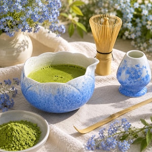 May include: A ceramic matcha bowl with a blue and white gradient finish, a bamboo whisk, a matching blue and white whisk holder, and a bamboo scoop. A small bowl contains green matcha powder, and the set is arranged on a textured fabric.