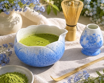 May include: A ceramic matcha bowl with a blue and white gradient finish, a bamboo whisk, a matching blue and white whisk holder, and a bamboo scoop. A small bowl contains green matcha powder, and the set is arranged on a textured fabric.