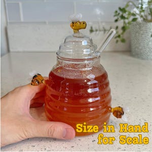 May include: A clear glass honey pot with a bee-shaped lid and two bee-shaped handles. The pot is filled with amber-colored honey and includes a glass honey dipper. The text "Size in Hand for Scale" is visible.