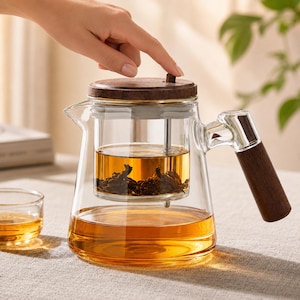 Pode incluir: Um bule de vidro transparente com tampa de madeira castanho-escura e uma pega lateral a condizer. O bule contém um infusor de vidro com folhas de chá submersas num chá de cor âmbar, com uma pequena chávena de vidro ao lado.
