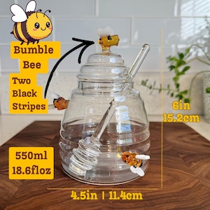 May include: A clear glass honey jar with a bee-shaped lid and two bee-shaped embellishments. The jar includes a glass honey dipper and holds 550ml or 18.6 fl oz. It measures 4.5 inches wide and 6 inches tall.