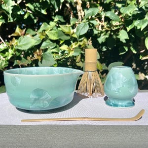 May include: A teal ceramic matcha bowl set with a whisk, a small tea cup, and a bamboo scoop. The bowl has a spout and a crystalline pattern. The whisk is made of bamboo. The tea cup and bowl are teal.