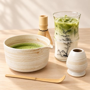 May include: A ceramic matcha bowl with a beige swirled glaze, a bamboo whisk, a bamboo scoop, a small ceramic whisk holder, and a tall glass filled with iced matcha latte featuring a black bamboo and mountain graphic print.
