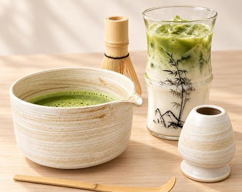 May include: A ceramic matcha bowl with a beige swirled glaze, a bamboo whisk, a bamboo scoop, a small ceramic whisk holder, and a tall glass filled with iced matcha latte featuring a black bamboo and mountain graphic print.