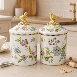 Puede incluir: Dos botes cilíndricos blancos con un colorido estampado floral de motivos botánicos en rosa, azul y verde. Cada bote tiene un borde dorado y una tapa a juego rematada con una figura decorativa de un pájaro.
