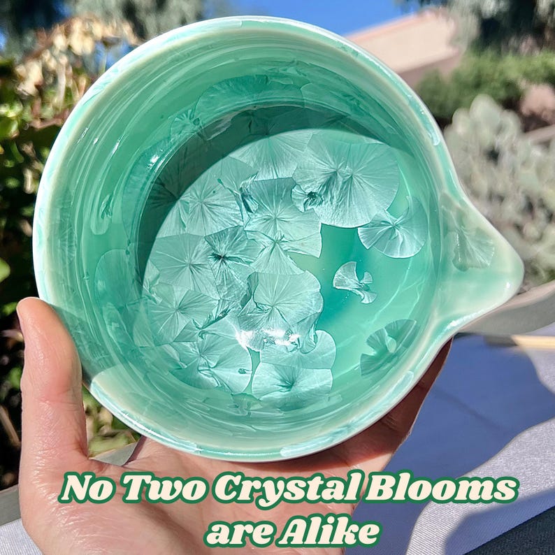May include: A round ceramic bowl with a glossy mint green glaze featuring white crystalline patterns that resemble flower blooms. The interior surface displays unique, radiating crystal formations. The text "No Two Crystal Blooms are Alike" is printed in green at the bottom.