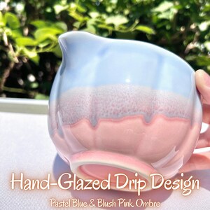 May include: A ceramic creamer pitcher featuring a hand-glazed drip design with a pastel blue and blush pink ombre finish. The pitcher has a rounded body, a small pouring spout, and a smooth, glossy surface texture throughout the exterior.