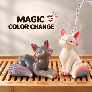 Puede incluir: Dos figuras de zorro con acabado brillante. Uno es gris oscuro con una cola en degradado rosa y azul, y el otro es blanco con acentos rosas en las orejas y una cola arcoíris. Agua cae sobre el zorro blanco. Texto: MAGIC COLOR CHANGE.