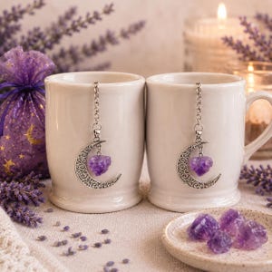 May include: Two white ceramic mugs each featuring a silver-toned metal tea infuser. The infusers have a decorative crescent moon charm with a small, rough-cut purple amethyst heart crystal suspended from a silver chain hanging inside the mugs.