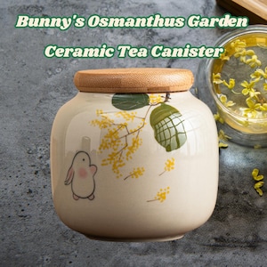 May include: Cream-colored ceramic tea canister with a bamboo lid. The canister is decorated with a bunny illustration and yellow floral sprigs. Text on the canister reads "Bunny's Osmanthus Garden Ceramic Tea Canister."