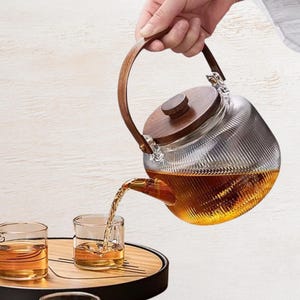 May include: A clear, ribbed glass teapot with a dark wood lid and a matching curved wooden handle. The teapot is tilted, pouring amber-colored tea into a small, clear glass cup. The design features a textured, vertical striped glass body.
