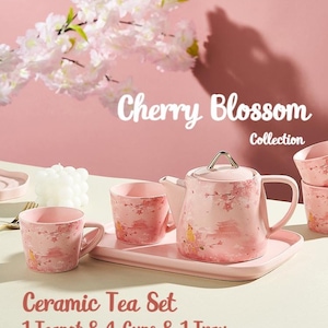 Op de afbeelding: Een roze keramische theeset met een theepot, vier kopjes en een dienblad. De set heeft een kersenbloesemontwerp in een roze en witte kleurstelling. De tekst "Cherry Blossom Collection" wordt boven de theeset weergegeven.