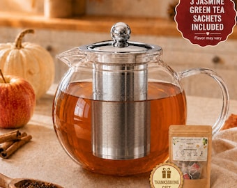 May include: A clear glass teapot with a stainless steel infuser filled with amber tea. A small brown paper pouch labelled Grape Jasmine Oolong is nearby. A red scalloped sign reads 3 JASMINE GREEN TEA SACHETS INCLUDED and a gold circular tag reads THANKSGIVING GIFT.