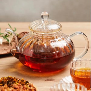 Op de afbeelding: Een heldere glazen theepot met een geribbeld ontwerp en een deksel. De theepot is gevuld met donkerbruine thee en een theezakje is zichtbaar in de pot. De theepot staat op een houten oppervlak.