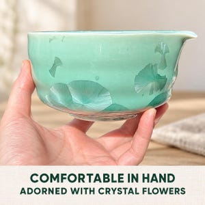 May include: A light mint green ceramic bowl featuring a crystalline glaze pattern that resembles delicate flower shapes. The bowl has a smooth, rounded body with a small pouring spout on the rim. Text on the image reads: COMFORTABLE IN HAND ADORNED WITH CRYSTAL FLOWERS.